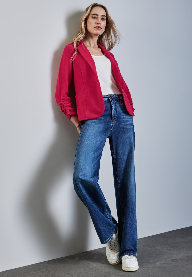 Femme en blazer rouge, haut blanc, jeans larges bleus et baskets blanches, appuyée contre un mur gris clair avec les mains dans les poches.