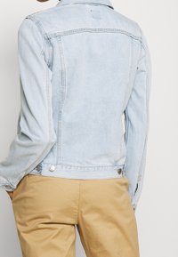 Světle modrá denimová bunda s klasickým střihem, která má límec, kovové knoflíky a viditelné švy. Nosená přes khaki kalhoty.