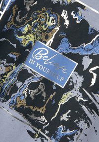 Schwarzer Stoff mit abstrakten Mustern in Blau, Grün und metallischem Gold. Ein rechteckiger blauer Bereich mit weißem Text trägt die Aufschrift "Glaube AN DICH SELBST."