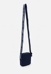 Bolso bandolera azul marino hecho de tela, con forma rectangular, correa ajustable con la marca "TOMMY JEANS" y cierre con cremallera.