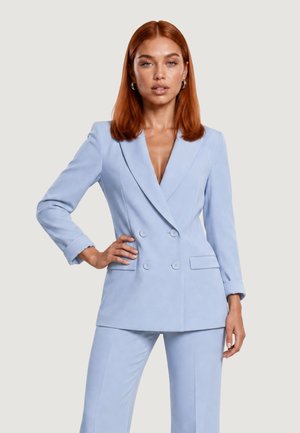 Femme aux cheveux roux portant un blazer bleu clair à double boutonnage et un pantalon assorti, debout avec une main sur la hanche devant un fond uni.