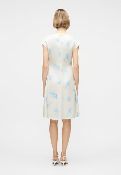 Γυναίκα που στέκεται με την πλάτη, φορώντας λευκό φόρεμα μέχρι το γόνατο με μπλε σχέδια tie-dye και λευκά σανδάλια με χαμηλό τακούνι και λουράκια στον αστράγαλο.