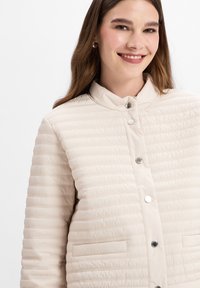 Beige gewatteerde jas met een staande kraag, voorkant met drukknoopsluiting en zijzakken. Kenmerkt zich door een textuurstof met horizontale stiksels.