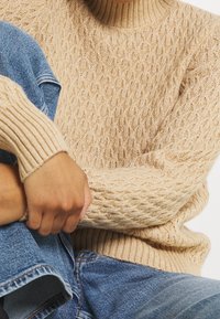 Person, der einen beigen Kabelstrickpullover und blaue Jeans trägt, sitzt mit einem Bein über das andere geschlagen und mit den Händen auf dem Knie.