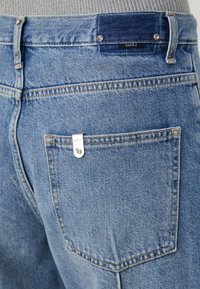Jeans de denim azul claro con un bolsillo trasero que presenta un acento metálico y una etiqueta de marca. Costuras distintivas y una textura ligeramente desgastada.