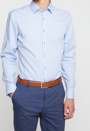 Homme portant une chemise bleu clair à manches longues rentrée dans un pantalon bleu avec une ceinture en cuir marron. Mains dans les poches.