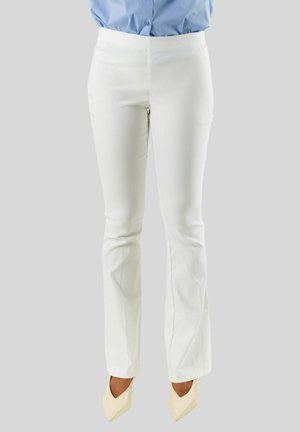 Donna che indossa pantaloni bianchi aderenti, scarpe beige con punta e una camicia azzurro chiaro infilata, su uno sfondo grigio uniforme.