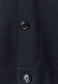 Marinblå stickad tröja med texturerad fabric, som har en rund svart knapp med blommönster och en liten knapphålsdetalj.