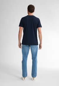 Marinblå t-shirt med rund hals och rosa-randiga ärmslut, kombinerad med ljusblå jeans och vita sneakers, sedd från baksidan.