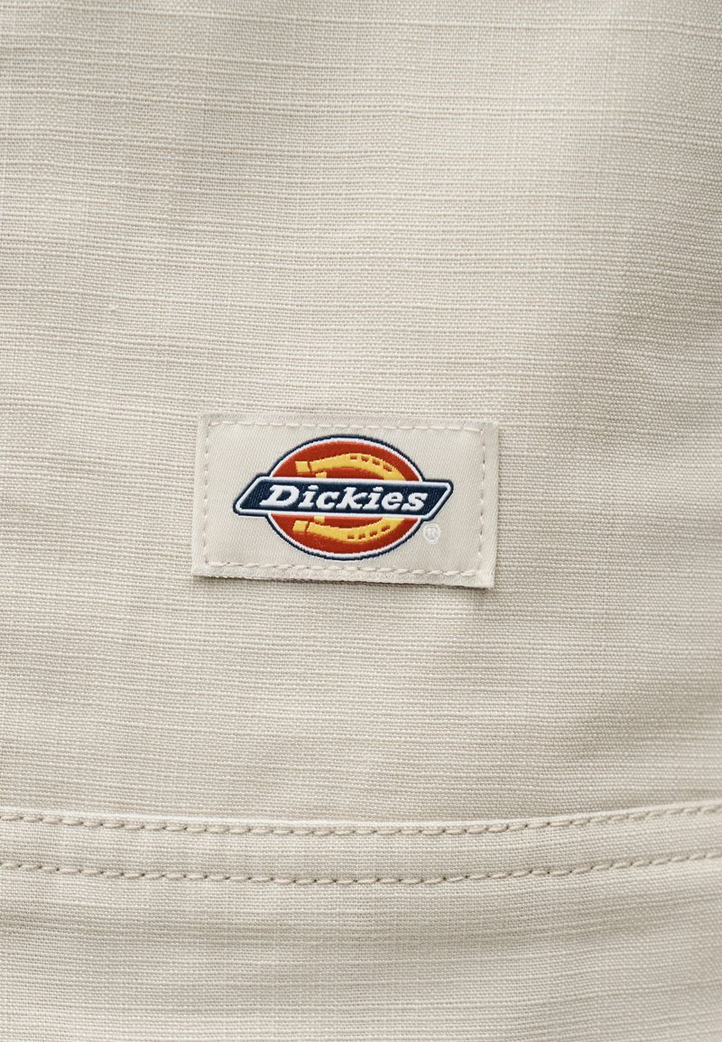 Beiger Stoff mit genähtem Saum und einem rechteckigen Etikett mit dem Dickies-Logo in Rot, Gelb, Weiß und Marineblau.