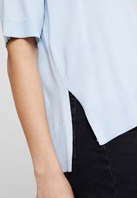 Ljusblå stickad tröja med korta ärmar, med sidospalter och ribbad nederkant, parat med mörka jeans. Slät struktur synlig.