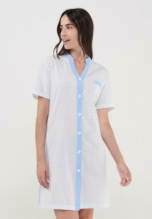 Mujer con vestido blanco de manga corta con botones, con lunares azules y ribete de cuadros, de pie frente a un fondo blanco liso.