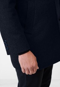 Marineblauer Wollmantel mit strukturierter Oberfläche, der über einen geknöpften Ärmelbund und eine aufgesetzte Tasche verfügt. Der Ärmel zeigt ein tailliertes Design und ein glattes Innenfutter.