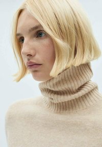 Beige turtleneck sweater i blød, ribbet strik, med høj krave og glat tekstur, båret af en person med blont hår.