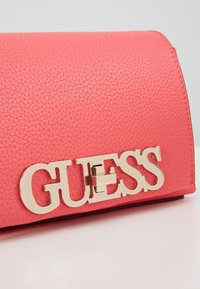 Borsa sintetica rosa con texture, caratterizzata dal logo "GUESS" in tonalità oro e forma rettangolare. Profilo liscio lungo i bordi.