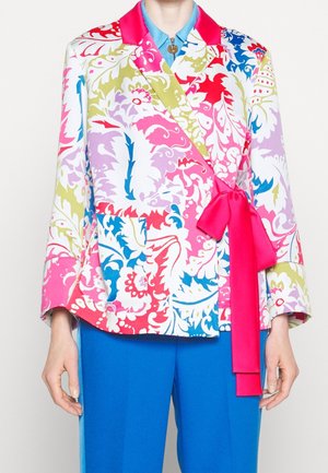Femme portant un blazer à motif floral multicolore avec un col et un nœud rose vif, associé à un pantalon bleu et une chemise bleue en dessous.
