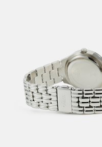 Zilveren metalen horloge met een gladde, samengestelde armbandontwerp. Heeft een ronde kast en een sluiting gemarkeerd met "GUESS." Gepolijste afwerking.
