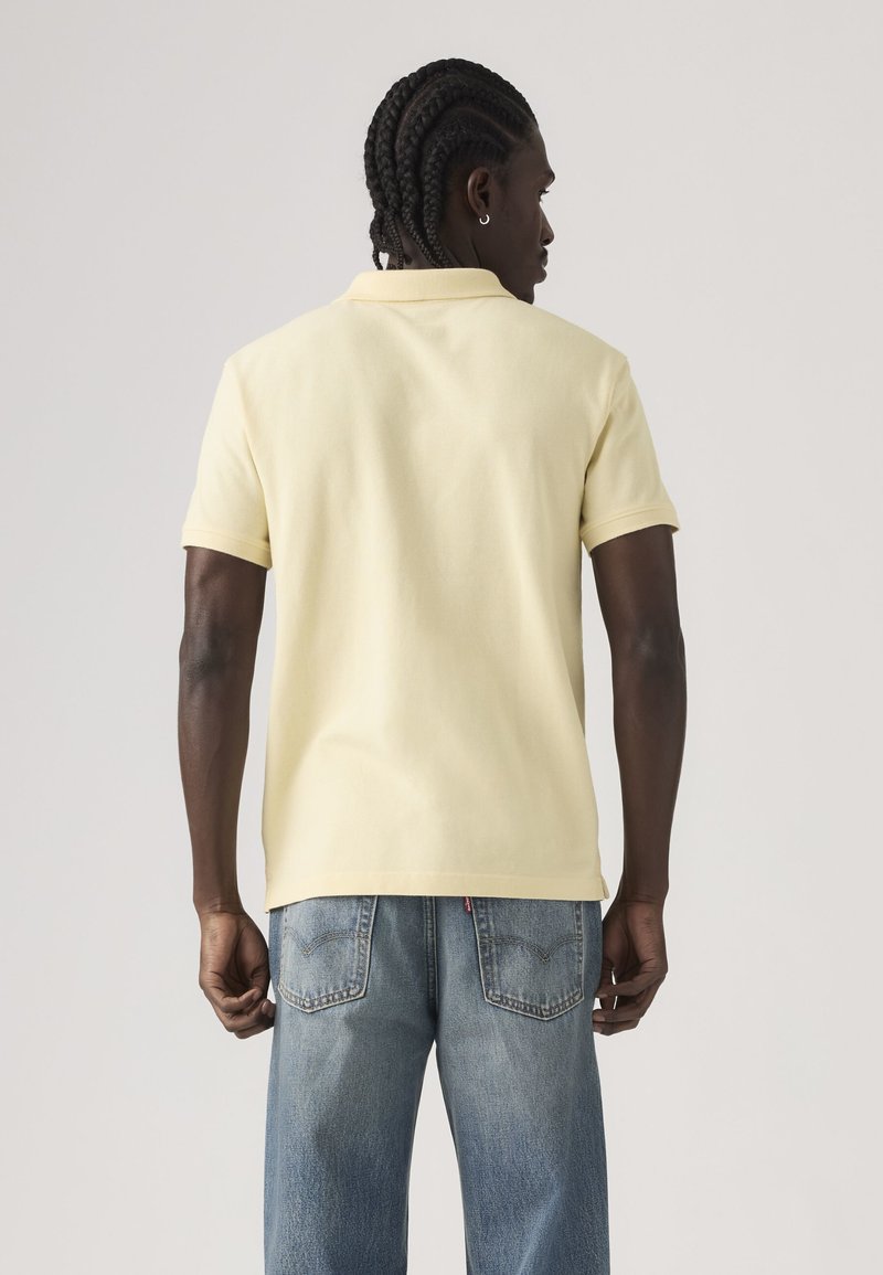 Homme aux cheveux tressés vu de dos, portant un polo à manches courtes jaune pâle et un jean en denim bleu clair.