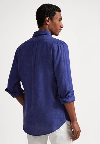 Μπλε λινό πουκάμισο με διπλωμένο γιακά και γυρισμένα μανίκια, με λεία υφή και καμπυλωτό τελείωμα, τοποθετημένο σε μοντέλο από πίσω.