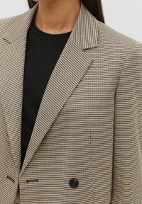 Brown and beige houndstooth blazer with peak lapels, featuring double button closure and textured fabric, layered over a black shirt.