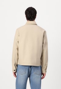 Beige jakke med krage og knappekanter, laget av slitesterk stoff. Har en boksformet design og sømdetaljer på baksiden, kombinert med lyseblå jeans.
