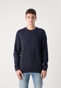 Tamsiai mėlynas ilgomis rankovėmis megztinis, pagamintas iš minkštos medžiagos, su apvaliu iškirpimu ir maža logotipu ant krūtinės, derinamas su šviesiai mėlynais džinsais.