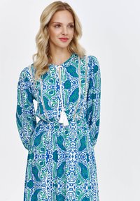 Sukienka w niebiesko-zielony wzór paisley, z długimi rękawami, okrągłym dekoltem i białymi frędzlami. Wykonana z lekkiej tkaniny z misternymi wzorami.