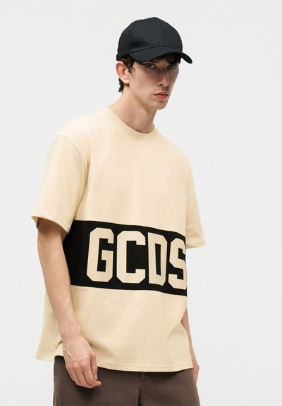 Νέος άνδρας που φοράει ένα μπεζ φαρδύ μπλουζάκι με μαύρη οριζόντια λωρίδα και γράμματα "GCDS", μαύρο καπέλο μπέιζμπολ και καφέ παντελόνι.