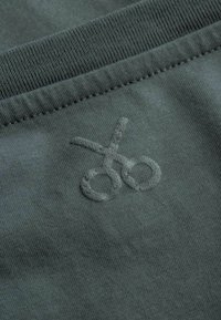 Donkergroene stof met een zachte textuur, met een subtiele geborduurde schaar logo in het midden, wat een minimalistisch ontwerp creëert.