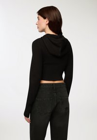 Felpa nera corta con orlo a costine e maniche lunghe, dotata di cappuccio; indossata con pantaloni in denim scuro che presentano due tasche posteriori.