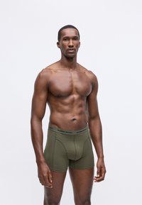 Uomo muscoloso in piedi, indossando boxer Björn Borg verde oliva, su uno sfondo chiaro e semplice, con lo sguardo rivolto in avanti.