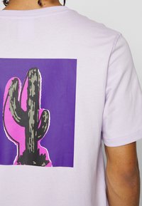 Osoba v světle fialovém tričku s černou a zelenou grafikou kaktusu uvnitř růžového a fialového čtverce na zádech.