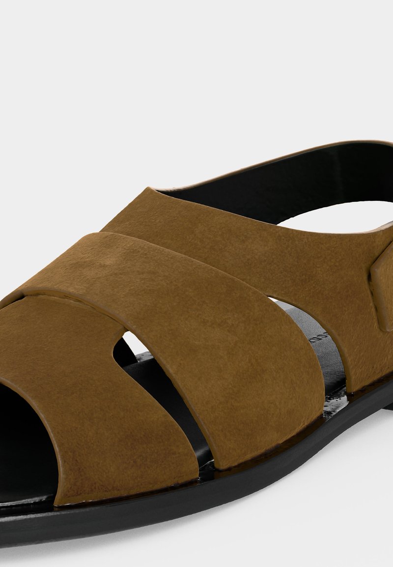 Sandalia marrón de cuero con puntera abierta, tiras anchas cruzadas y suela negra sobre fondo blanco.