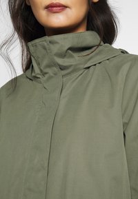 Svetle olivově zelená bunda s kapucí vyrobená z hladkého materiálu, s vysokým límcem a subtilními švy pro elegantní design.