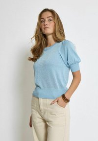 Hellblaues kurzärmliges Sweatshirt mit strukturiertem Muster, gerundetem Ausschnitt und geripptem Saum. Kombiniert mit beigen, hoch sitzenden Hosen.