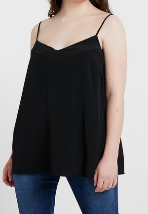 Woman wearing loose black spaghetti strap top and blue jeans, standing against a plain background.