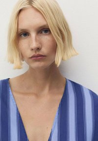 Păr blond, ochi albaștri și pistrui. Modelul poartă un top cu decupaj adânc în V, cu dungi verticale albastre și albastre deschise pe un material moale. Design simplu.