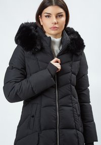 Chaqueta acolchada negra con cierre frontal, diseño acolchado y capucha forrada de piel sintética. Cuenta con bolsillos laterales y puños ajustados. Hecha de material resistente al agua.