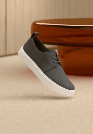 Donkergrijze casual sneaker met witte zool, zwevend boven een beige tapijt, met daarachter een glanzende bruine salontafel.