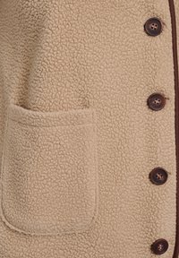 Beige fleecejakke med en lomme og mørkebrunne knapper. Stoffets tekstur er blød og struktureret med en let fuzzy overflade.