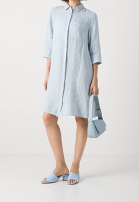 Lichtblauw gestreepte jurk met driekwartmouwen, knoopsluiting en een borstzakje. Gecombineerd met lichtblauwe blokhak sandalen en een handtas.