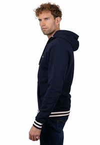 Sudadera con capucha azul marino que incluye un bolsillo frontal, puños y dobladillo acanalados con rayas blancas. Tela suave con una textura lisa.