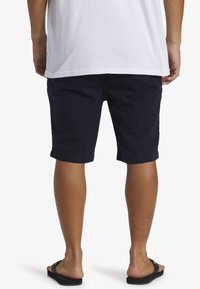 Pantalones cortos de algodón azul marino, hasta la rodilla, con un corte relajado y bajos doblados. Llevados con chanclas negras sobre pies descalzos.