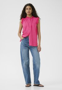 Canottiera senza maniche fucsia con colletto arricciato, chiusura con bottoni, abbinata a jeans blu a vita alta e ciabatte nere. Tessuto liscio e leggero.