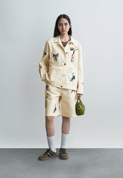 Μπεζ βαμβακερό μπουφάν με κεντημένα σχέδια ζώων, συνδυασμένο με ταιριαστά σορτς. Κρατά μια μικρή πράσινη τσάντα. Φορά λευκές κάλτσες και σκούρα αθλητικά παπούτσια.