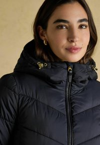 Czarna kurtka puchowa z wysokim kołnierzem, z zamkiem błyskawicznym i złotymi elementami wykończenia. Teksturowany, pikowany wzór zapewniający izolację.