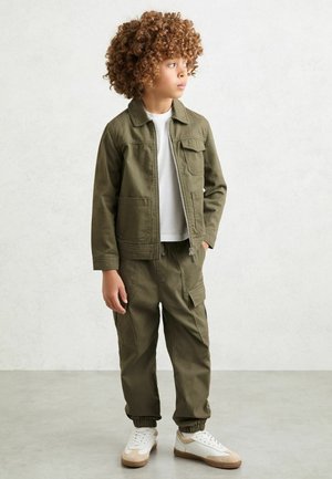 Lapsi, jolla on kiharat hiukset ja yllään oliivinvihreä takki ja sivutaskuilla varustetut housut, valkoinen t-paita ja valkoiset lenkkarit, katse kääntyen sivulle.