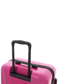 Hard-shell pink suitcase with a glossy finish, featuring a black telescopic handle and a textured grip area. Zipper closure along the edge.