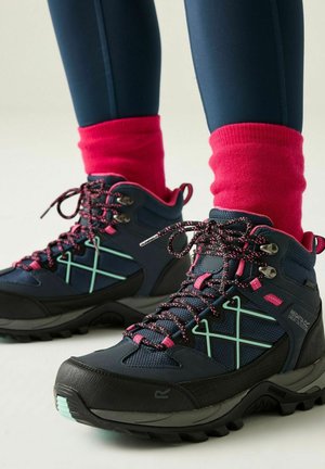 Bottes de randonnée bleu marine avec lacets et accents roses, portées avec des chaussettes rouges vives et des leggings bleu marine, présentées sur un fond clair.