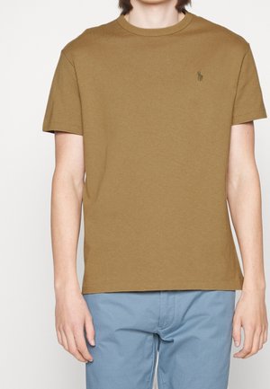 Persoon in een bruin T-shirt met korte mouwen en een geborduurd logo op de borst, met lichtblauwe broek, staand voor een witte achtergrond.